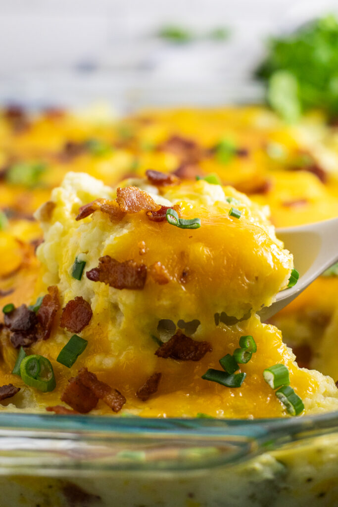 Side view of a silicone spoon scooping twice baked potato casserole topped with melted cheddar cheese, crumbled bacon, and sliced green onions.