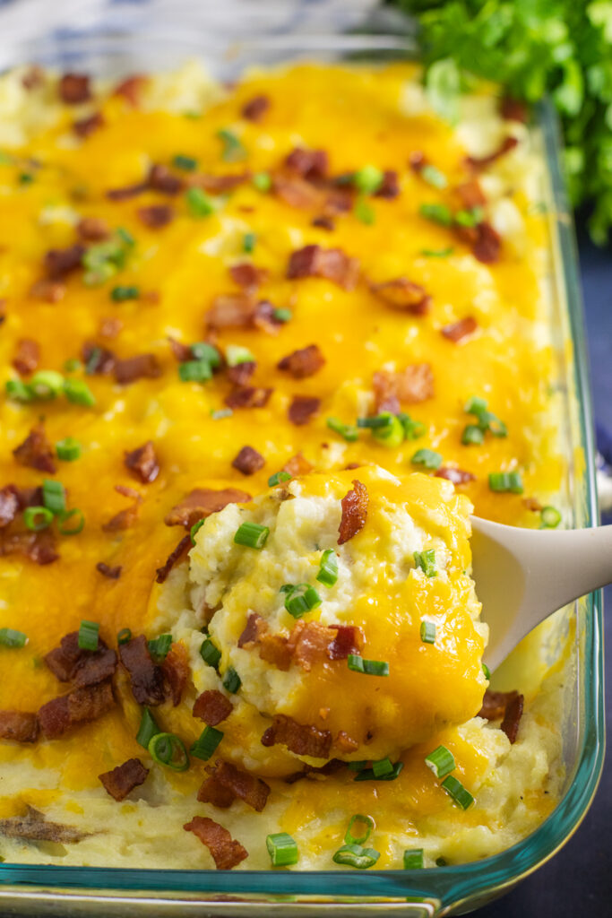 Overhead view of A silicone spoon scooping creamy twice baked potato casserole topped with melted cheddar cheese, crumbled bacon, and sliced green onions.
