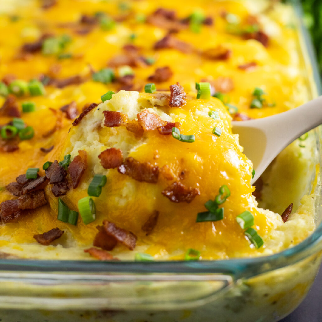A silicone spoon scooping twice baked potato casserole topped with melted cheddar cheese, crumbled bacon, and sliced green onions.