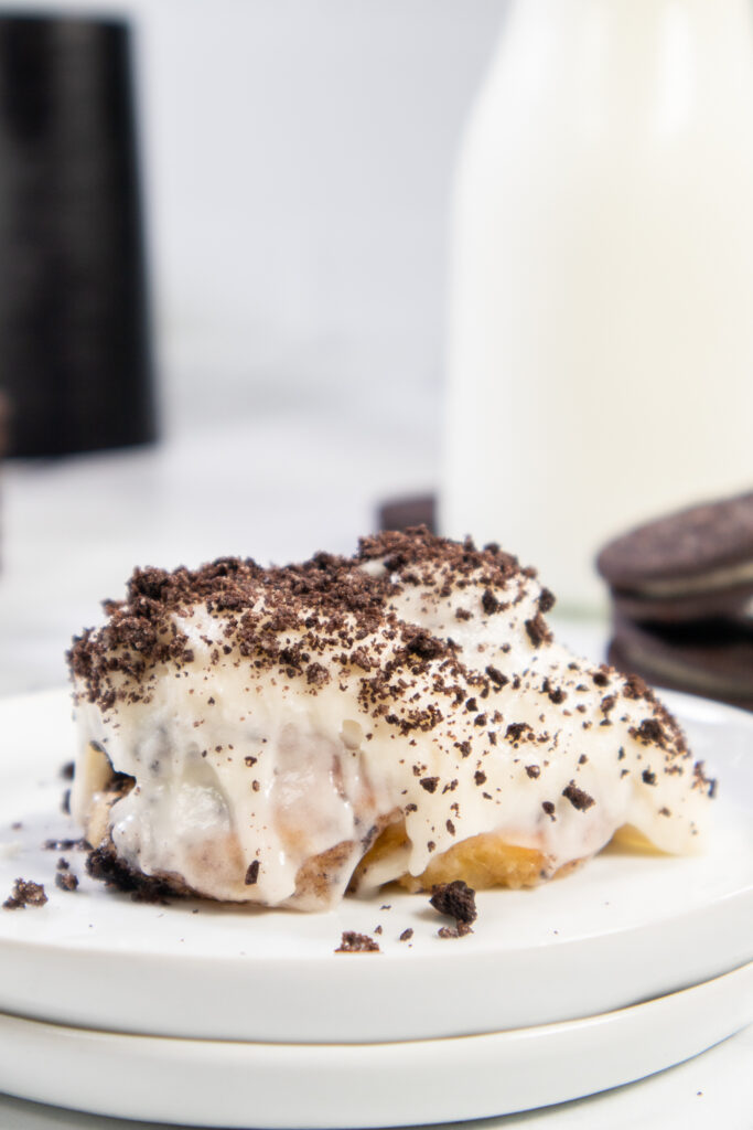 An Air Fryer Oreo Cinnamon roll on a small white plate topped with cream cheese frosting and crushed Oreo pieces.