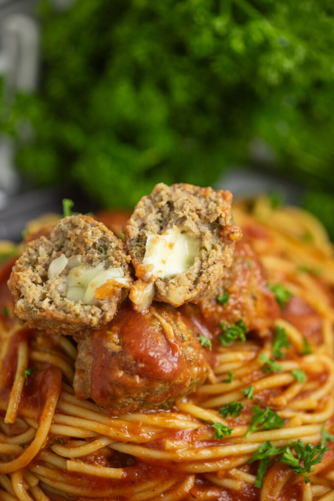 A slow cooker mozzarella stuffed meatball cut open to see the cheese between the two halves on top of plate of spaghetti topped with freshly chopped parsley.
