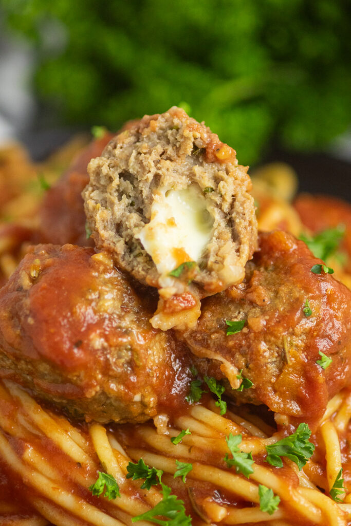 A close up of slow cooker mozzarella stuffed meatball cut open to see the cheese on top of 2 more meatballs on top of plate of spaghetti topped with freshly chopped parsley.
