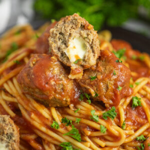 A slow cooker mozzarella stuffed meatball cut open to see the cheese on top of 2 more meatballs on top of plate of spaghetti topped with freshly chopped parsley.