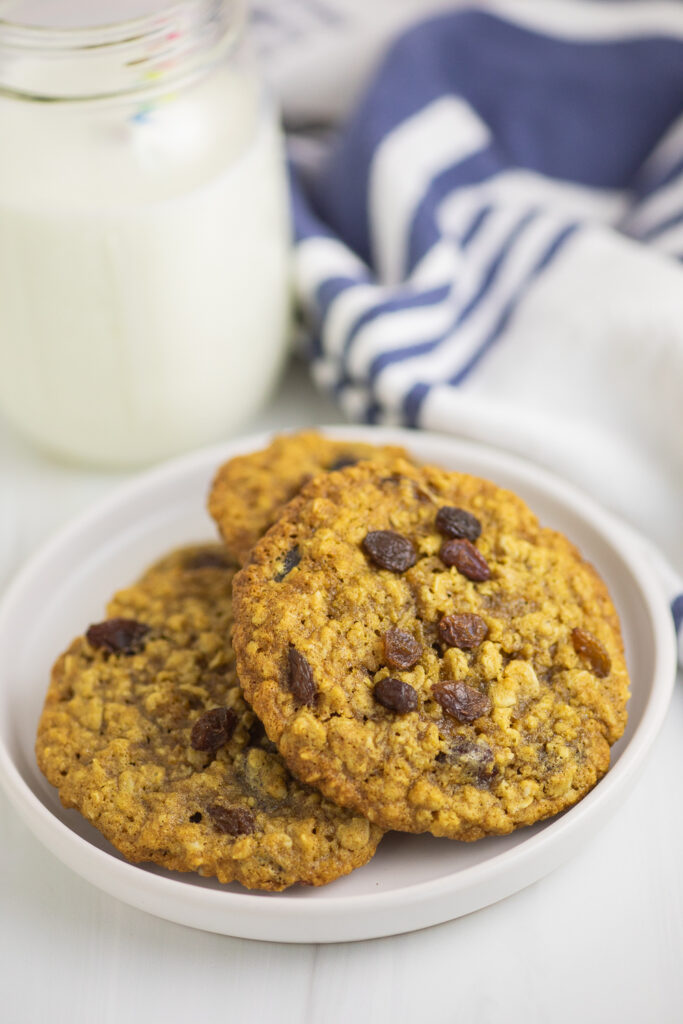 Oatmeal raisin cookies on a plate with a glass of milk and a linen napkin behind.