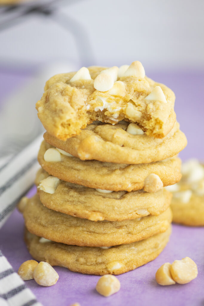A stack of white chocolate macadamia nut cookies. A bite is taken out of the top one and they are surrounded by macadamia nuts and a linen napkin.