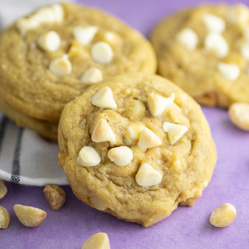 A white chocolate macadamia nut cookie showing the top surrounded by macadamia nuts with two more cookies and a linen napkin behind.
