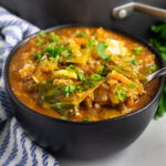 A black matte bowl full of cabbage roll soup topped with fresh chopped parsley with a spoon taking a scoop of the soup.