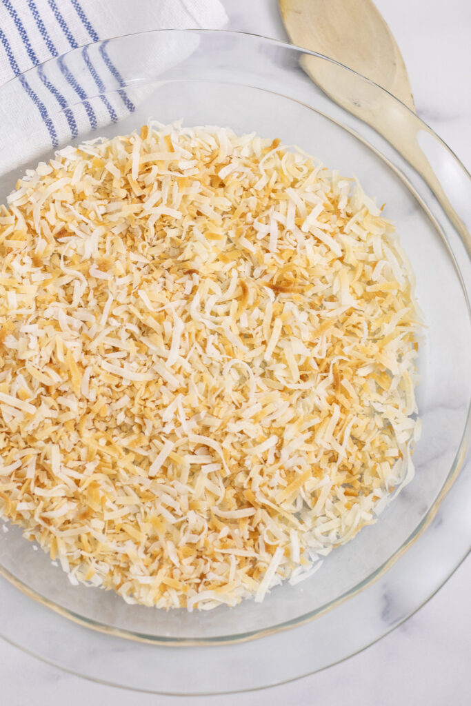 Overhead view of toasted coconut in a round glass pie dish next to a linen napkin and a thin wooden spoon.