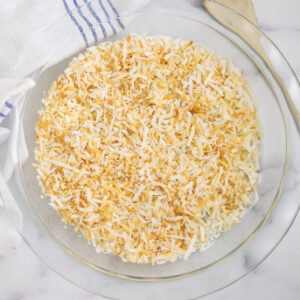 A round glass dish with toasted coconut. The dish is surrounded by a linen napkin and a thin wooden spoon.