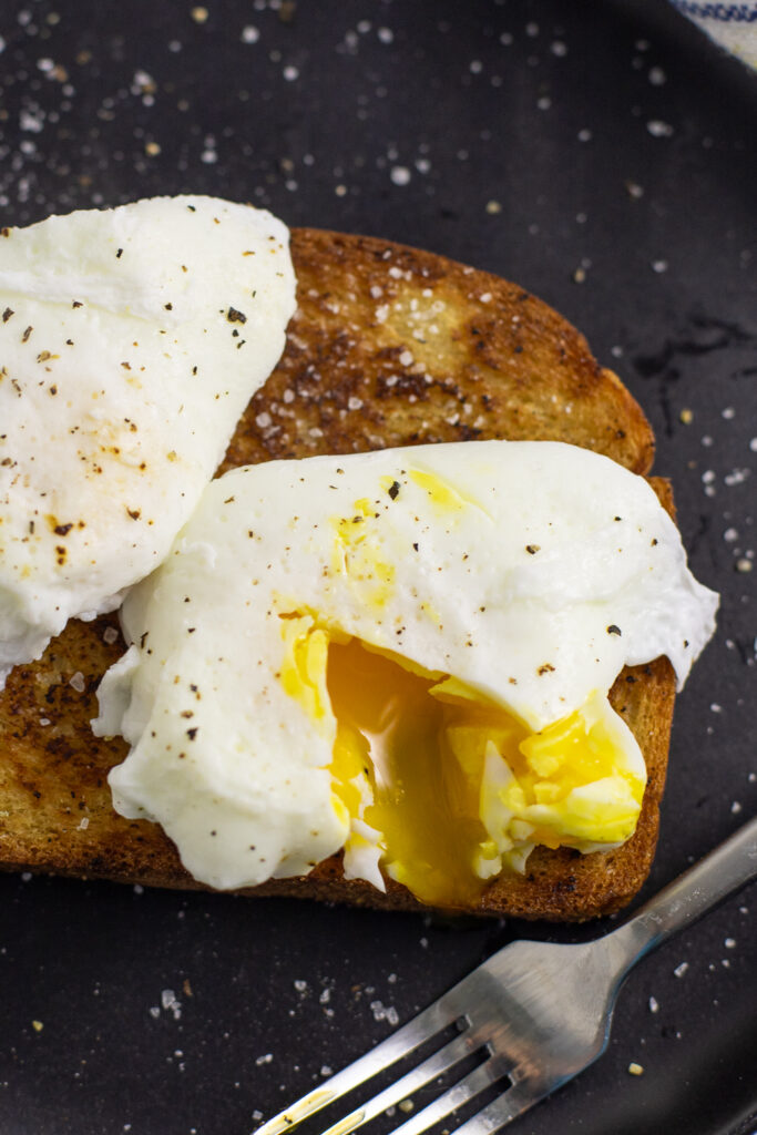 Overhead view of a poached egg topped with black pepper, cut into so that the yolk is running out on a piece of toast.