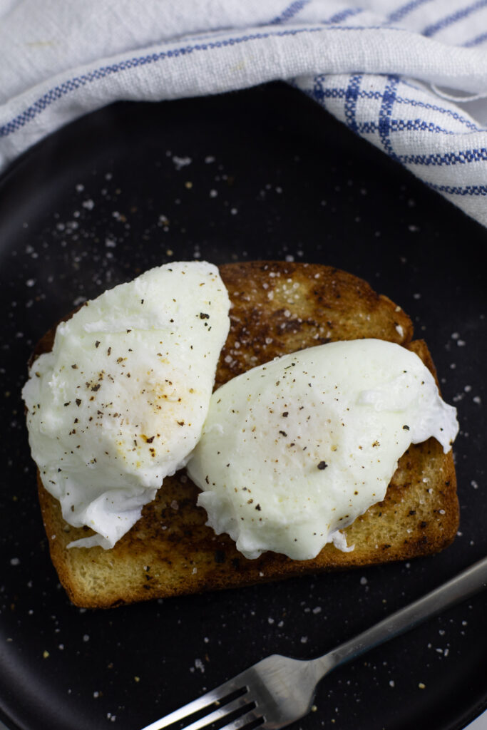 Two poached eggs topped with black pepper on a piece of toast on a black matte plate surrounded by a linen napkin.