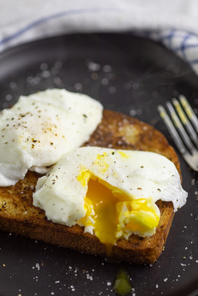 Poached egg topped with black pepper, cut into so that the yolk is running out with another egg behind on a piece of toast.