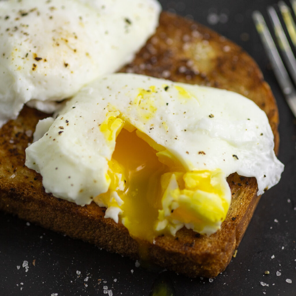 Poached egg topped with black pepper, cut into so that the yolk is running out on a piece of toast.