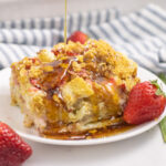 A piece of strawberry cream cheese French toast casserole on a small white plate next to a fresh strawberry with maple syrup drizzling on top.