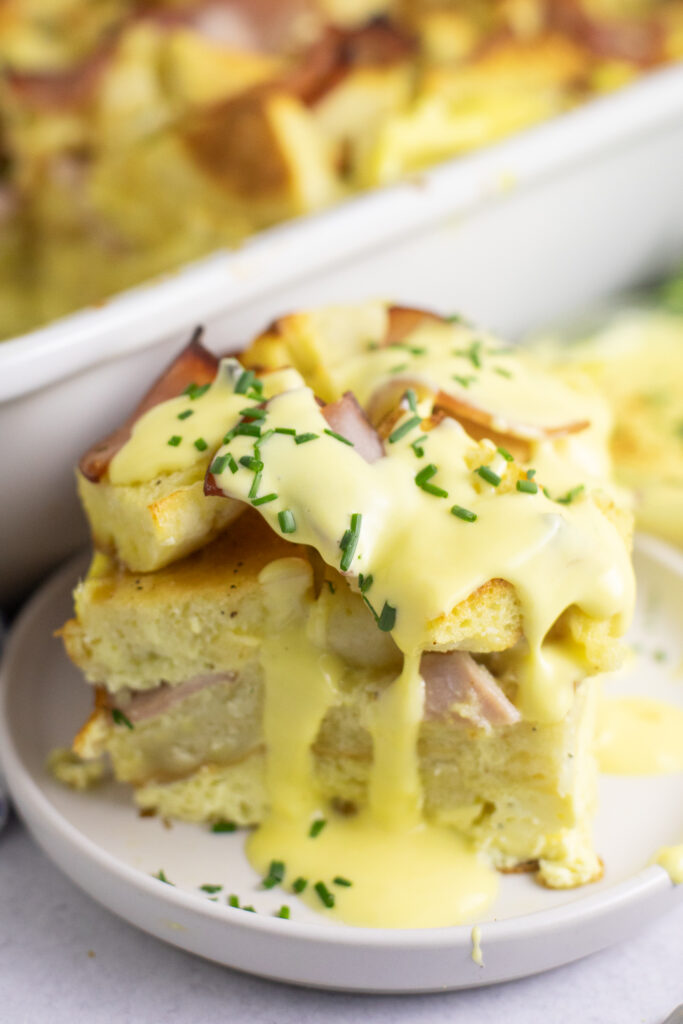 A piece of eggs benedict casserole on a small plate with hollandaise dripping down the sides and topped with fresh chopped chives.