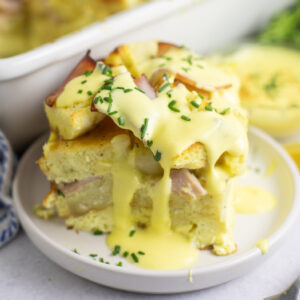 A piece of eggs benedict casserole on a small plate with hollandaise dripping down the sides and topped with fresh chopped chives.