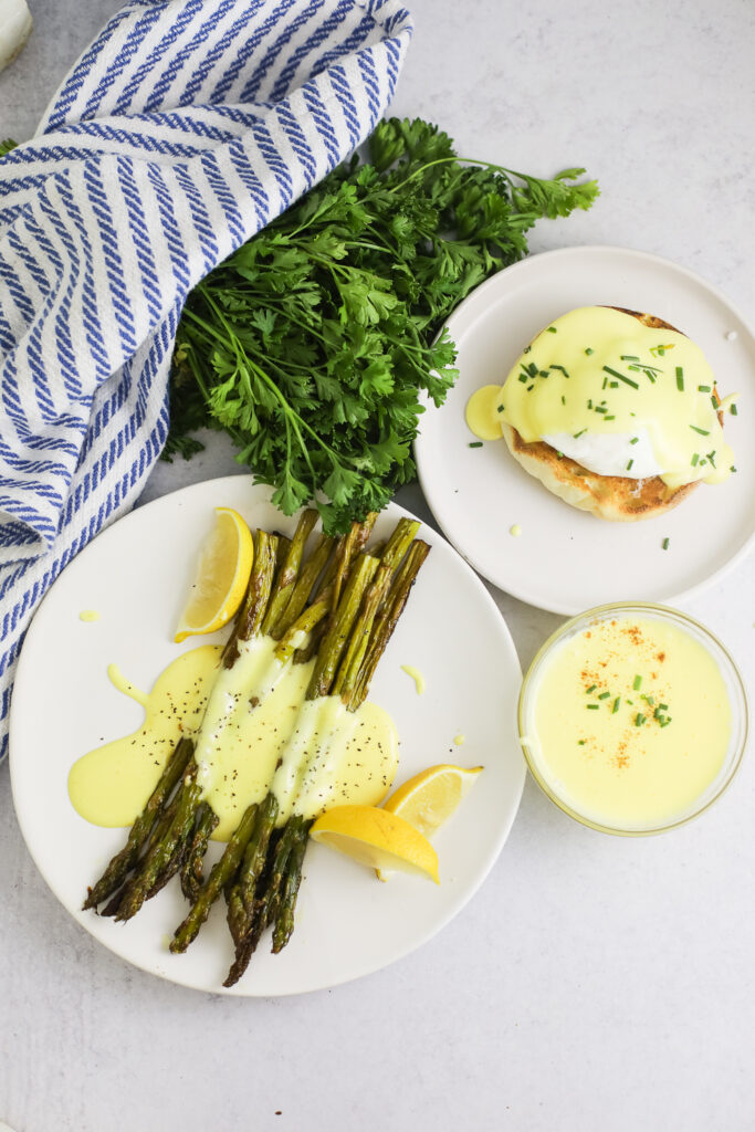Overhead view of a plate of asparagus with lemon wedges with hollandaise sauce on top, a poached egg on an English muffin topped with hollandaise sauce, a small glass bowl of hollandaise sauce, a fresh bunch of parsley, and a linen napkin.