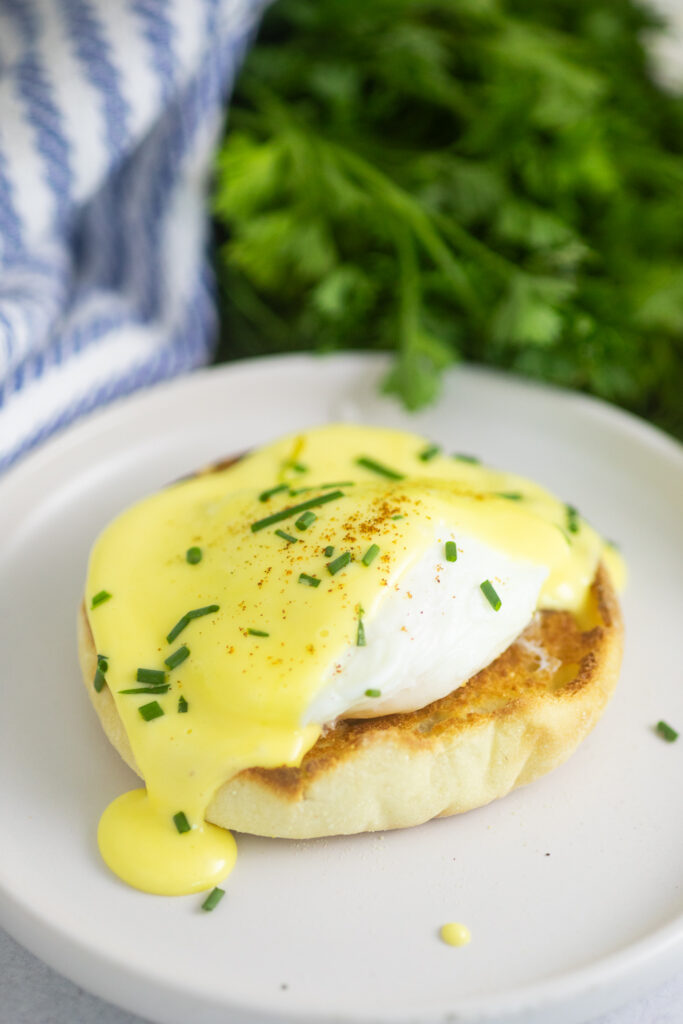 A poached egg on half of an English muffin topped with hollandaise sauce dripping down the side and fresh chives on top with a bunch of parsley and a linen napkin behind.