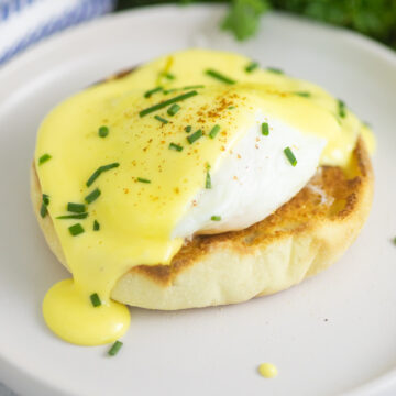 A poached egg on half of an English muffin topped with hollandaise sauce dripping down the side and fresh chives on top.