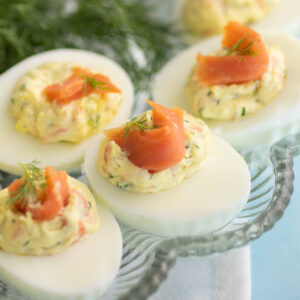 Smoked salmon deviled eggs topped with a piece of cold smoked salmon and a sprig of fresh dill on a deviled egg plate next to fresh dill.