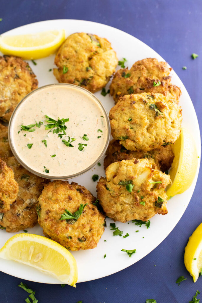 Air fryer crab cakes on a large plate with lemon wedges surrounding a small glass bowl of remoulade sauce topped with freshly chopped parsley.