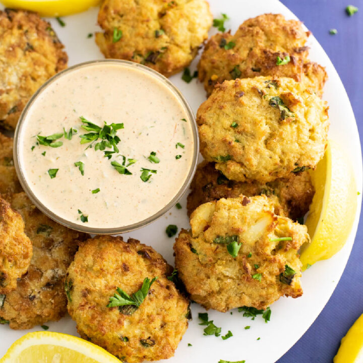 Air fryer crab cakes on a large plate surrounding a small glass bowl of remoulade sauce topped with freshly chopped parsley.