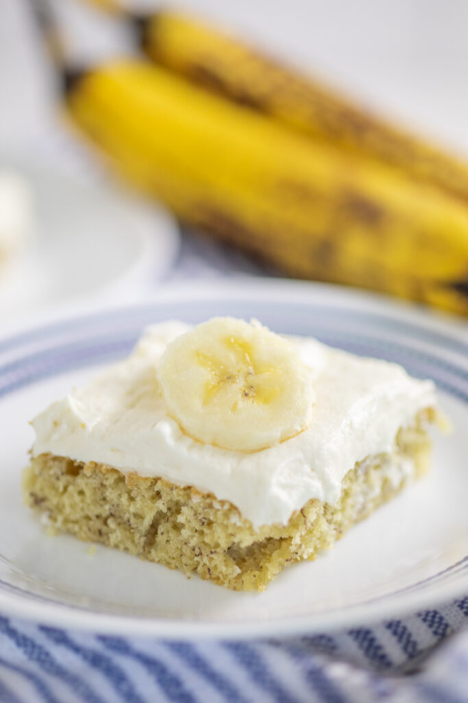 A banana bar with cream cheese frosting topped with a fresh banana slice on a small plate with fresh bananas behind it.