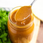 A spoon taking a scoop out of a glass jar full of homemade red enchilada sauce next to a bunch of fresh cilantro.