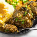 Instant Pot Salisbury steak patty with a bite taken out on a fork topped with gravy and freshly chopped parsley on a matte black plate next to mashed potatoes topped with gravy.