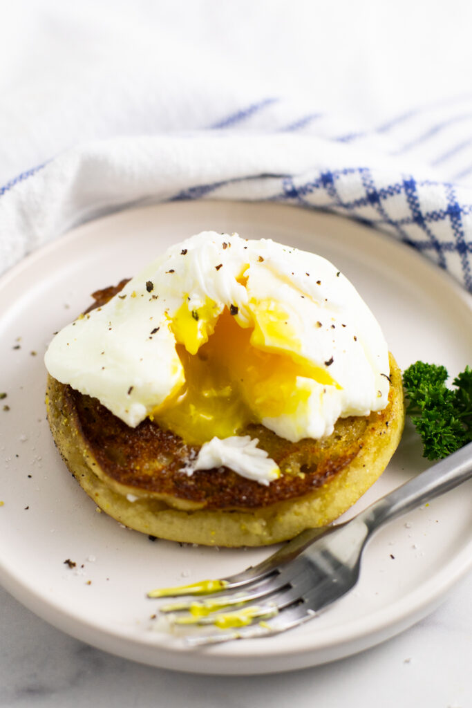 Microwave poached egg topped with black pepper cut open with yolk running out on half of an English muffin next to a metal fork on a small plate next to a linen napkin.