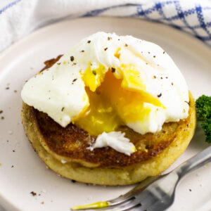 Microwave poached egg topped with black pepper cut open with yolk running out on half of an English muffin next to a metal fork.