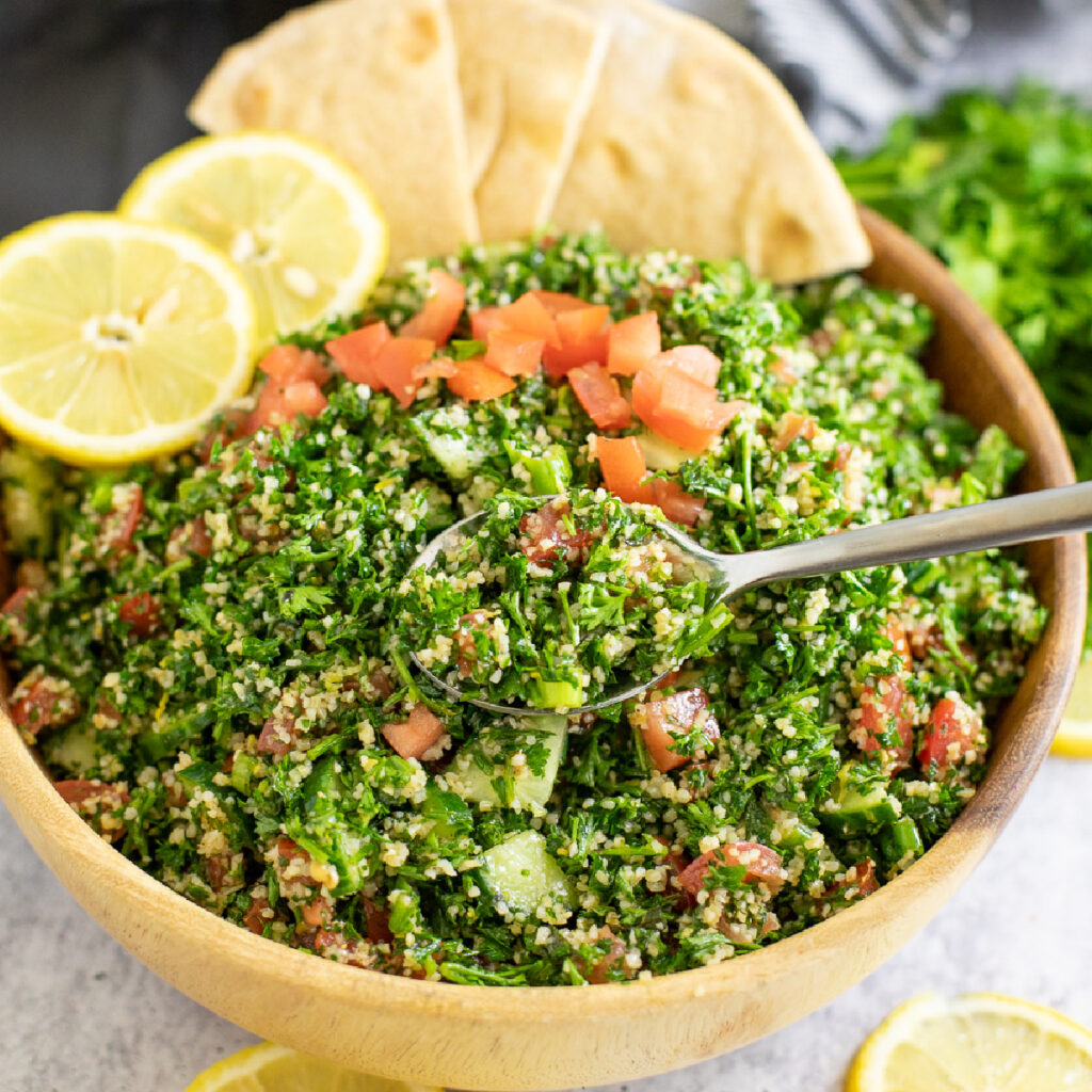 Tabouli Salad in a wooden bowl topped with fresh tomatoes, two lemon slices, wedges of pita, with a spoon scooping out a bite.