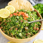 Tabouli Salad in a wooden bowl topped with fresh tomatoes, two lemon slices, wedges of pita, with a spoon scooping out a bite.