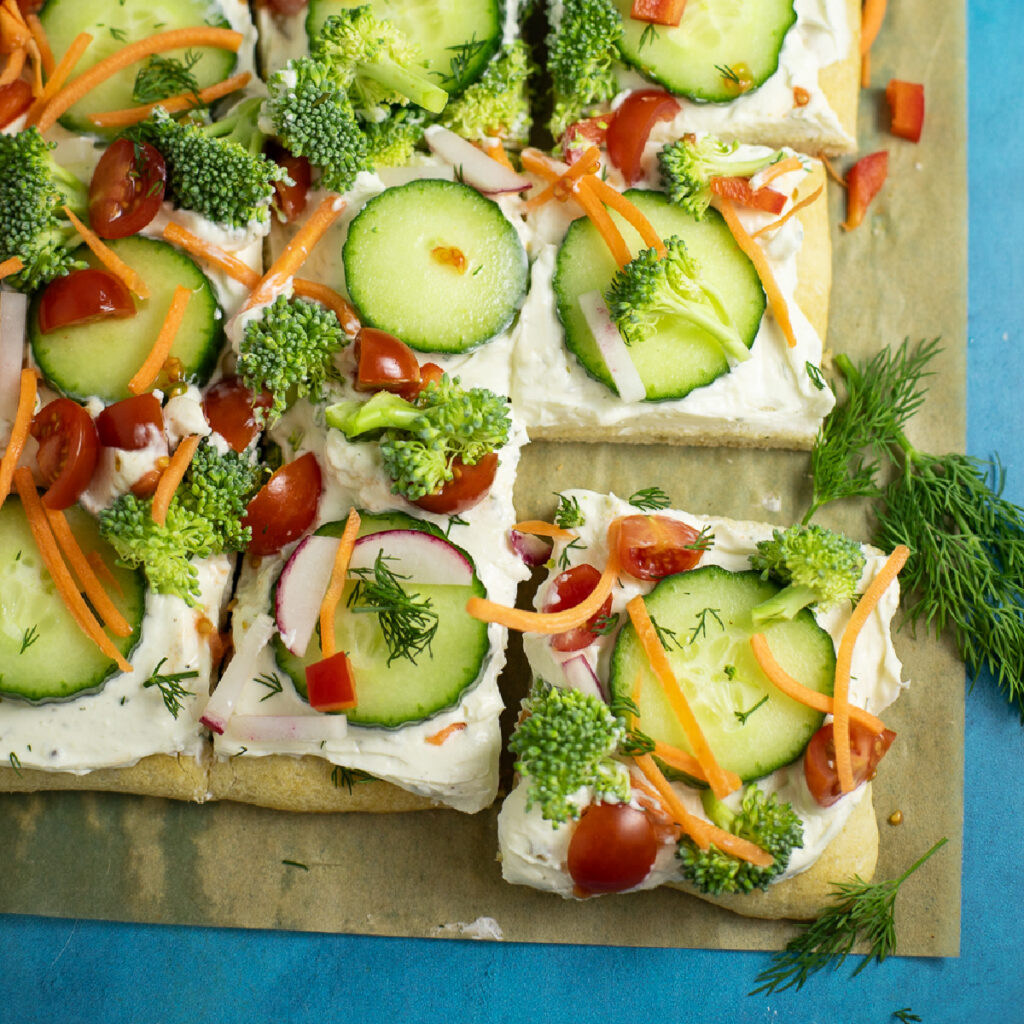 Overhead view of a cold veggie pizza topped with veggies like cucumber slices, shredded carrots, cherry tomatoes, fresh dill, and more, sitting on a parchment paper with a slice pulled out away from the rest.