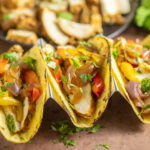 Three tacos filled with Instant Pot chicken fajita filling with chicken, bell peppers, and onions and topped with freshly chopped cilantro on a rustic background.