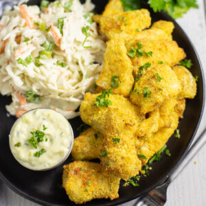 Air Fryer Catfish Nuggets topped with parsley on a black plate next to coleslaw and a small bowl of tartar sauce.
