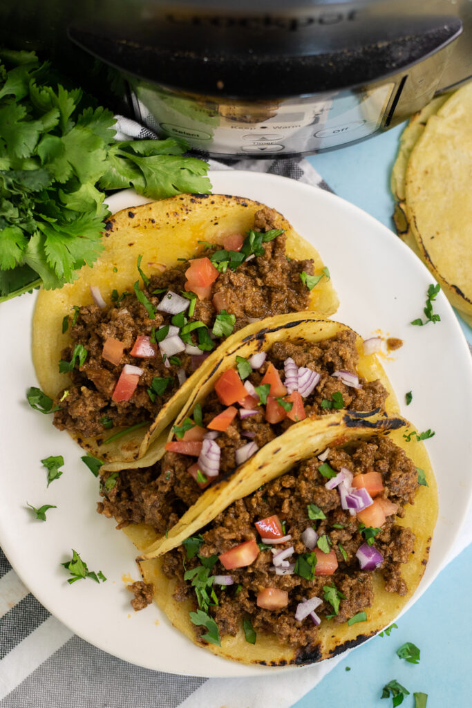Overhead view of three ground beef tacos filled with crockpot taco meat on a small white plate topped with pico de gallo next to a bunch of fresh cilantro and a slow cooker.