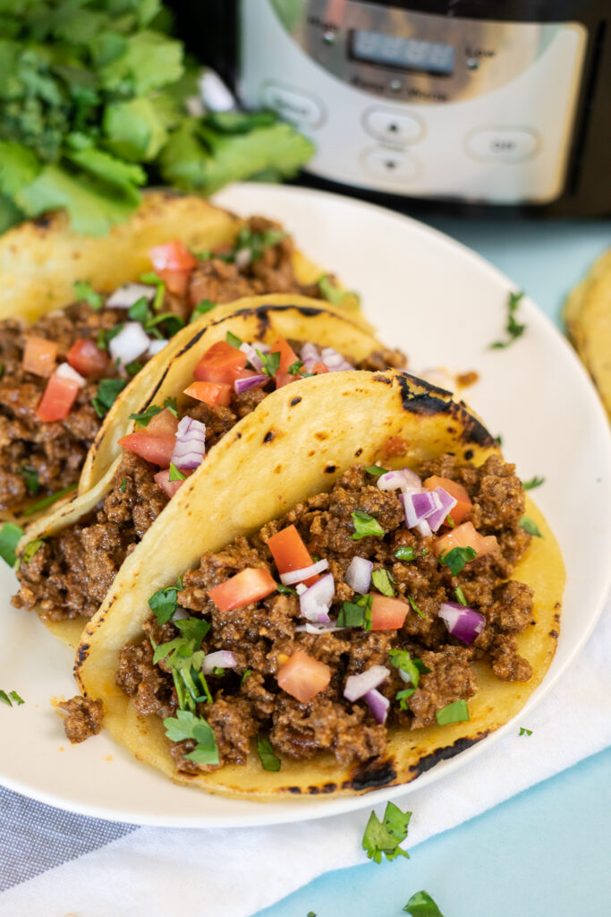 Close up of a ground beef taco in a corn tortilla filled with crockpot taco meat topped with pico de gallo on a plate with two other tacos with a slow cooker and a bunch of fresh cilantro behind.