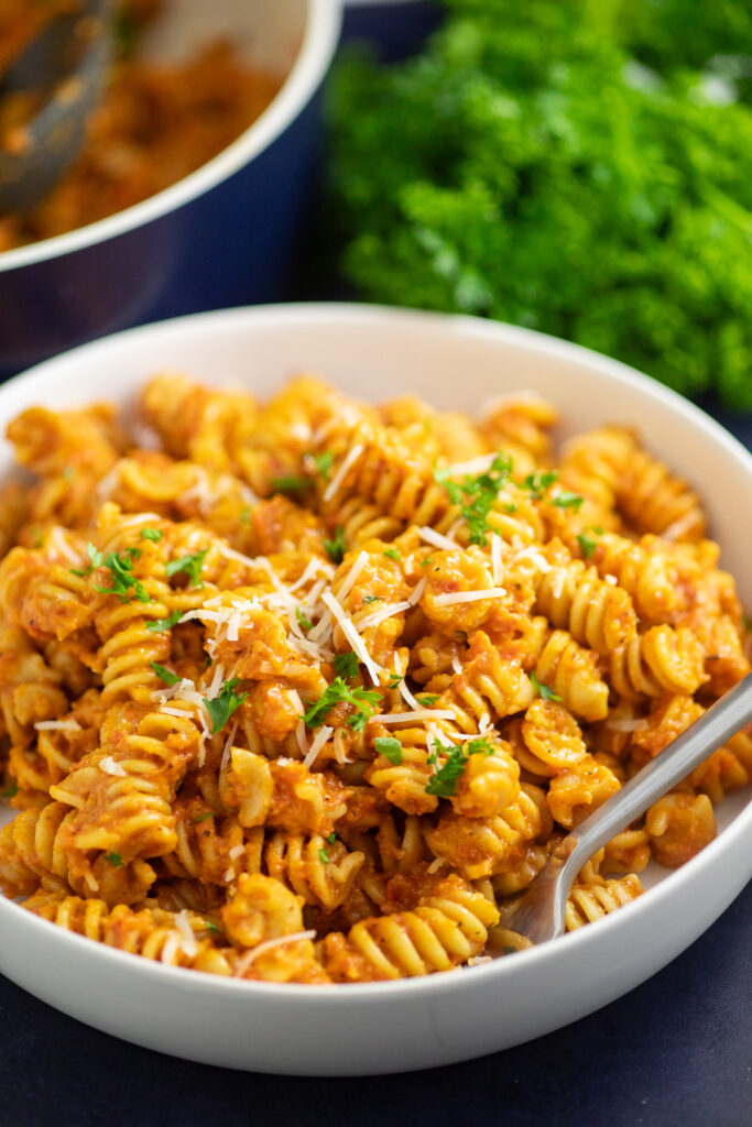 Romesco Pasta in a bowl with a fork topped with fresh parsley and manchego cheese with parsley and a navy blue skillet behind.