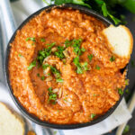 Overhead view of homemade romesco sauce in a small black bowl topped with fresh parsley with a baguette slice sticking out on the side.