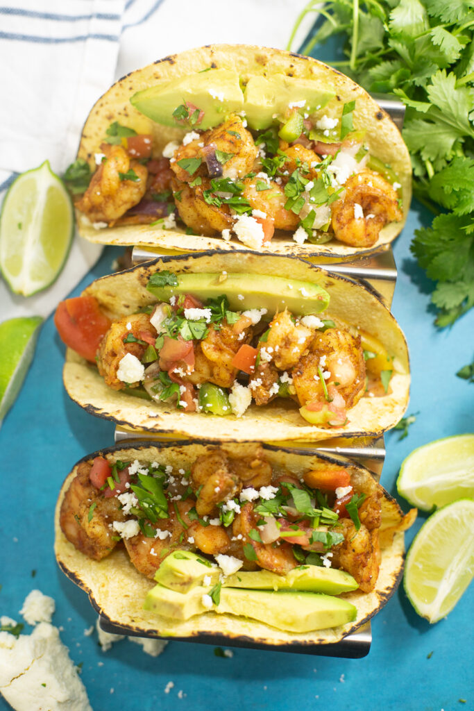 Overhead view of three sheet pan shrimp fajitas in corn tortillas topped with fresh cilantro, queso fresco, and avocado slices, and surrounded by lime wedges, a bunch of fresh cilantro, and a linen napkin.