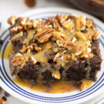 A piece of turtle poke cake topped with pecans and caramel sauce on a small plate.