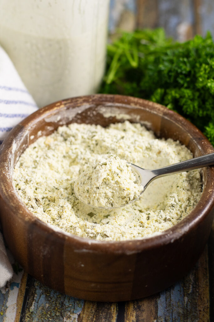 Homemade ranch seasoning in a wooden bowl with a spoon holding a scoop with a bunch of fresh parsley and a jar of ranch dressing behind.