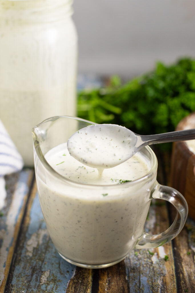 Homemade ranch dressing made from homemade ranch seasoning in a small glass dish with a spoon pouring a scoop out, a jar full of the same ranch and a bunch of parsley behind.