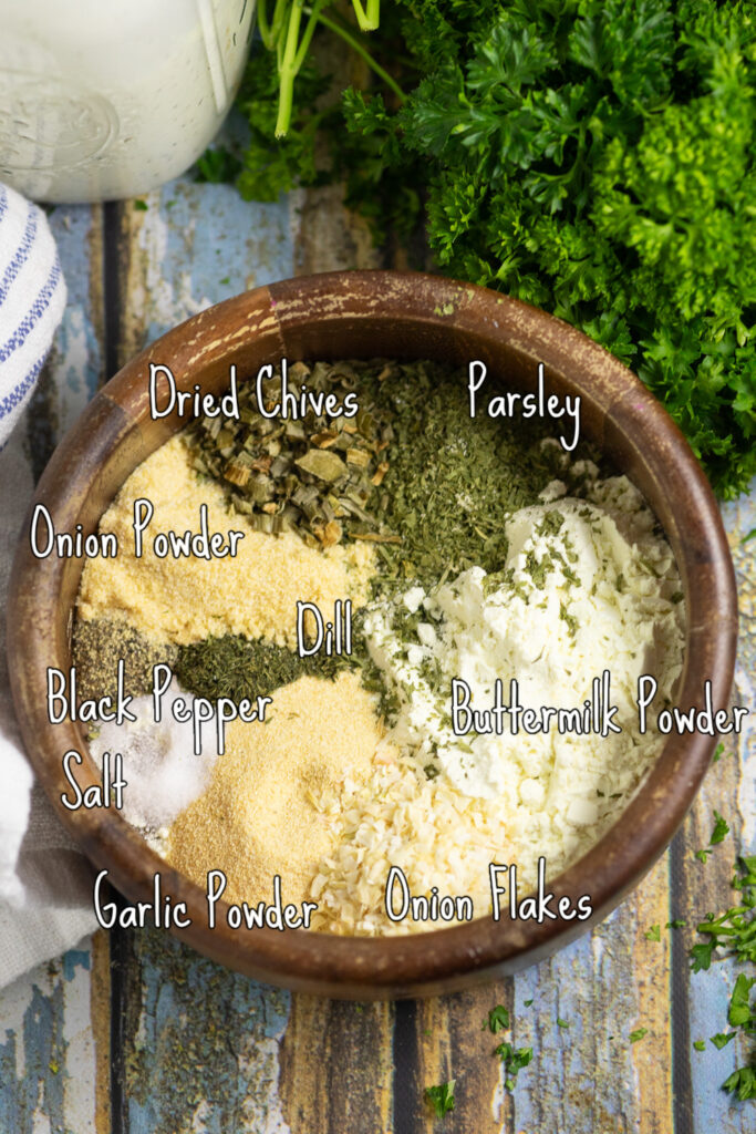 A wooden bowl with all of the ingredients to make homemade ranch seasoning such as parsley, buttermilk powder, onion flakes, garlic powder, salt, black pepper, dill, onion powder, and dried chives on a rustic wooden background next to a bunch of fresh parsley.