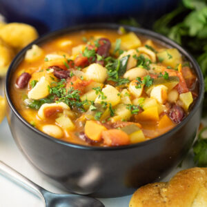 Homemade Olive Garden Minestrone Soup in a black matte bowl next to a spoon and breadstick in front of a large blue pot, more breadsticks, and a bunch of fresh parsley.