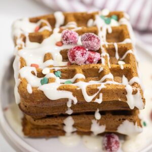 Two waffles stacked on top of each other drizzled with vanilla glaze and three sugared cranberries on a small ceramic plate.