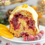 Orange cranberry bundt cake topped with orange glaze and an orange peel curl, sitting on a small white plate next to two orange slices and sugared cranberries.