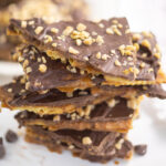 A stack of saltine toffee topped with toffee bits with chocolate chips next to it, and a plate of toffee behind.