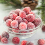 Sugared Cranberries in a small glass bowl surrounded by more sugared cranberries on a white marble counter with a pine branch and pinecone in background.
